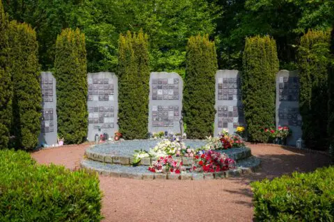 Gedenkpanelen tussen hoge groene hagen rond een ronde gedenkplaats met bloemen.