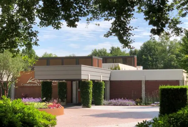 Voorgevel en ingang van Crematorium Kranenburg, omringd door bomen, hagen en bloeiende beplanting.